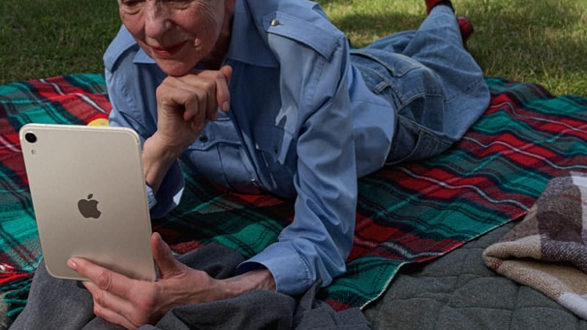 A person lying on a blanket in a park reading on the new iPad mini