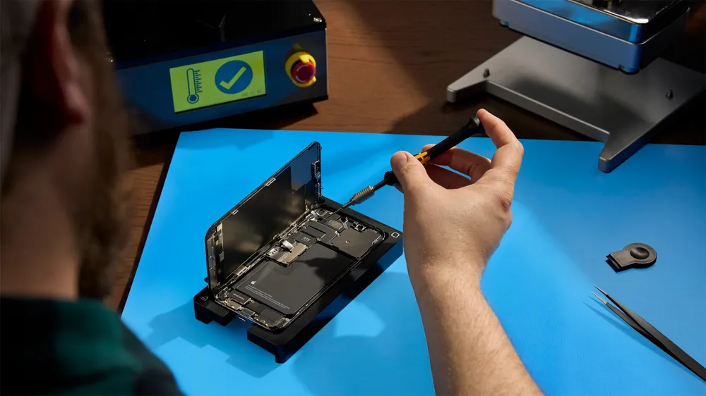 A technician works on an open iPhone held in Apple's repair tray, with a repair press in the background